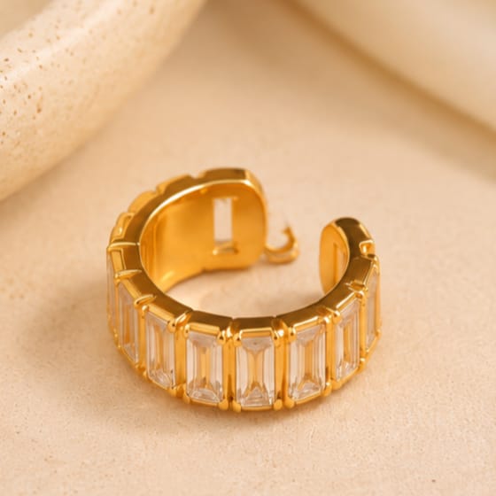 A gold-toned open cuff ring featuring a row of clear baguette-cut crystals, displayed on a textured beige surface.
