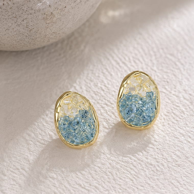A pair of gold-framed stud earrings with textured blue and white resin inlays, displayed on a textured white surface next to a ceramic bowl.