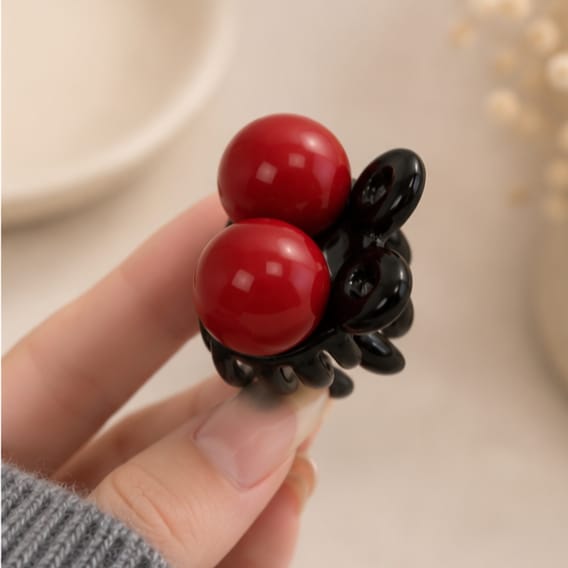 A close-up of a hand holding a black claw hair clip decorated with two glossy red spherical beads, set against a soft, warm beige background.
