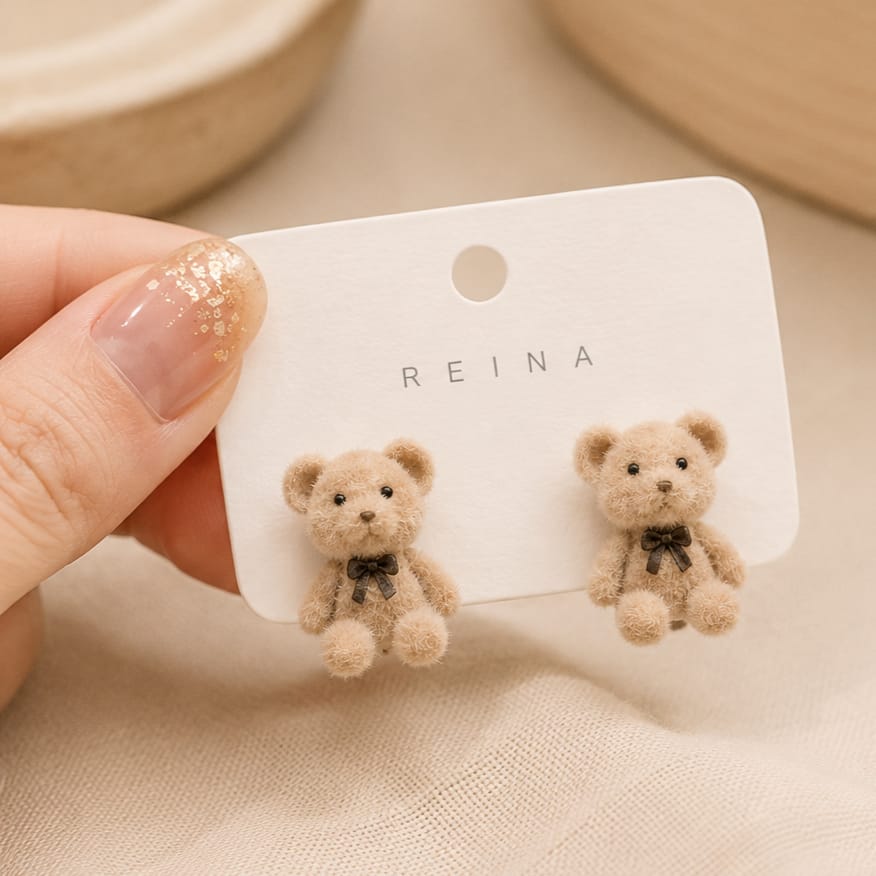 A pair of small teddy bear stud earrings on a white display card held by a hand. The bears are light beige with black bow ties, and the card is printed with the brand name REINA.
