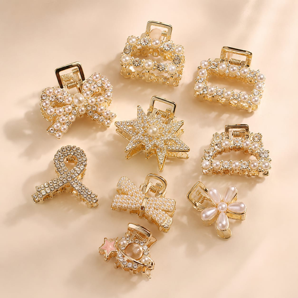 Nine decorative hair claw clips arranged on a soft beige background. The clips feature gold-tone metal settings adorned with rhinestones, pearls, and some with pastel pink faux pearls.