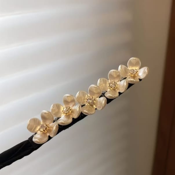 A black hair accessory with five glossy cream flower charms, each detailed with a small gold center, shown against a soft-focus background of white window blinds.