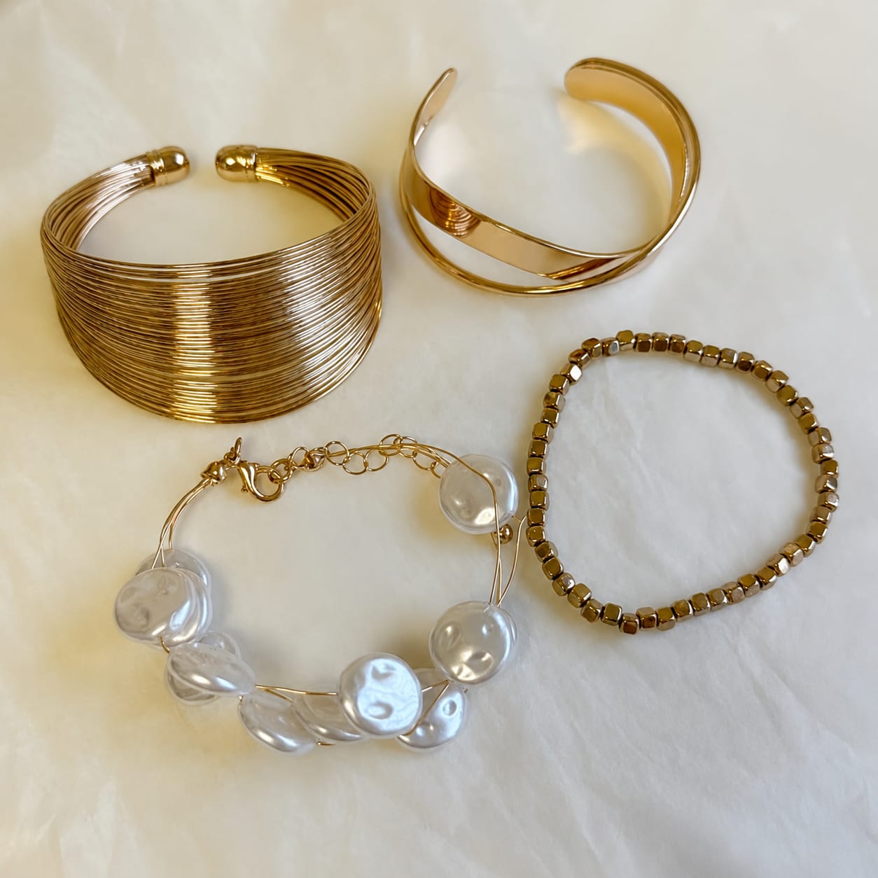 Four jewelry pieces arranged on a white fabric background: two gold-toned cuff bracelets, one gold-toned beaded bracelet, and one bracelet with irregular white pearl-like beads on a gold chain.