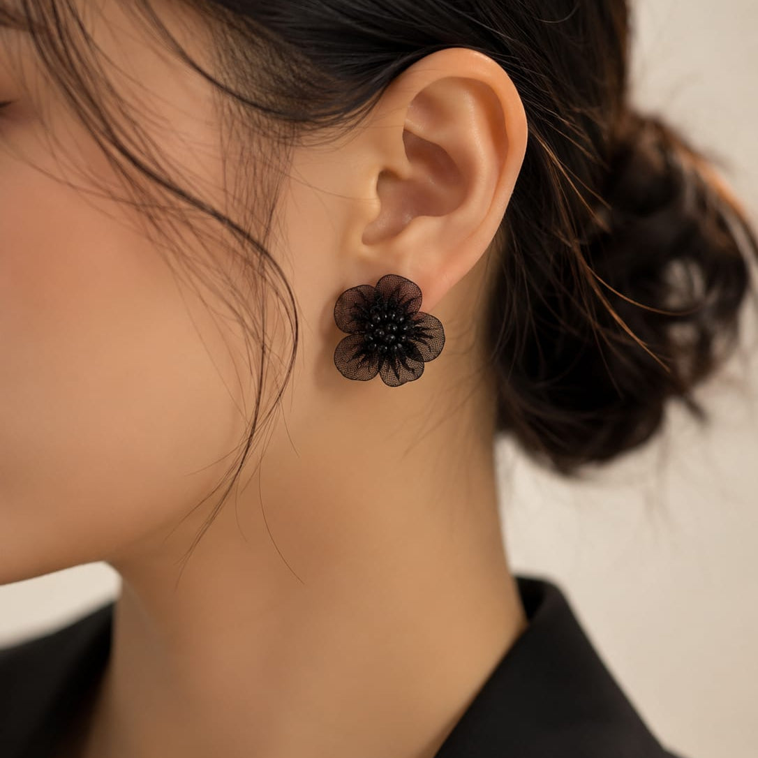 A close-up of a model's ear wearing a black flower-shaped stud earring, with her dark hair tied back and a black top visible against a light neutral background.