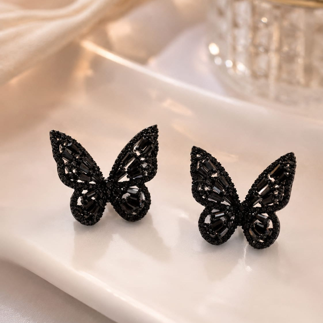 A pair of black butterfly-shaped stud earrings with intricate textured detailing, displayed on a white ceramic tray.