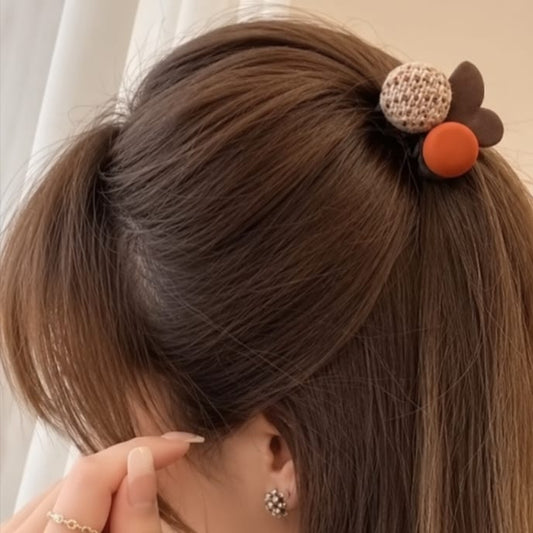 A close-up of a person's brown hair styled in a half-up ponytail with a decorative hair tie featuring a knitted beige bun, a dark brown flower, and an orange accent.