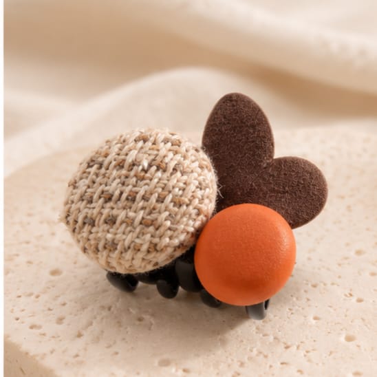 A small decorative hair claw clip shaped like a ladybug, featuring a beige woven fabric body, a brown felt wing, and an orange felt belly, shown on a soft cream textured surface.