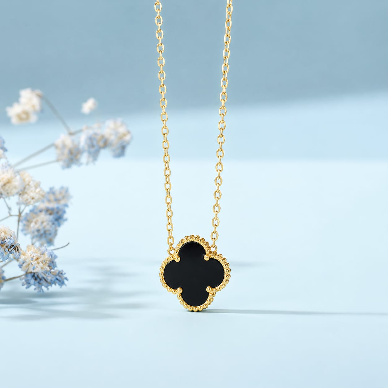 A gold chain necklace with a black clover pendant, displayed against a soft light blue background with delicate dried flowers on the left.