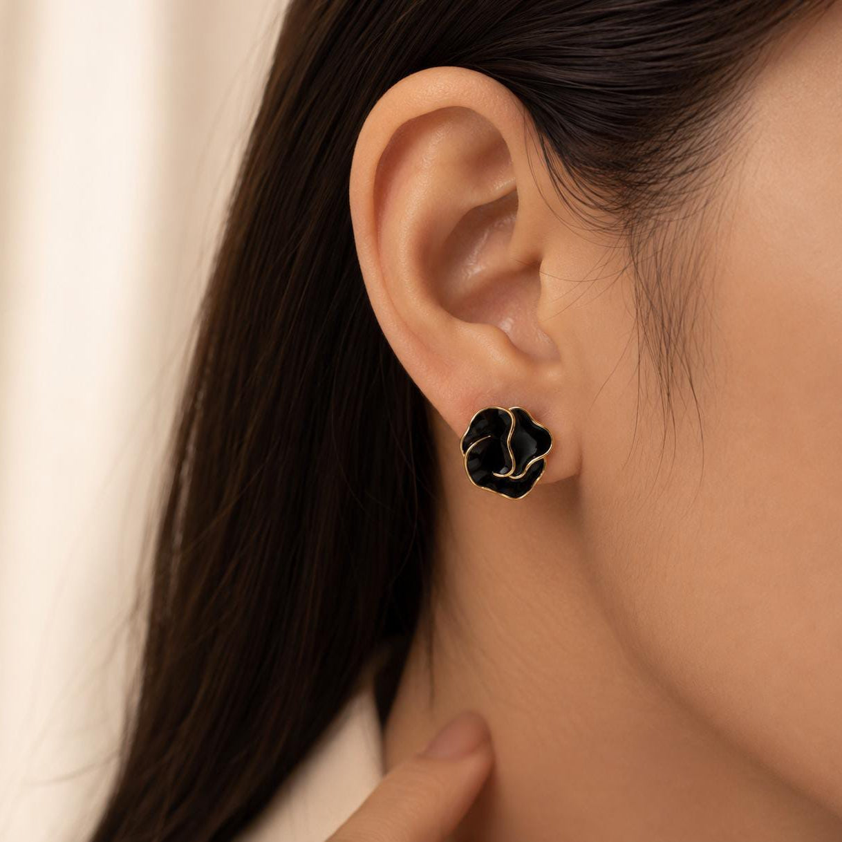 A close-up of a person's ear wearing a black flower-shaped stud earring with gold-toned metal accents, against a soft cream background.