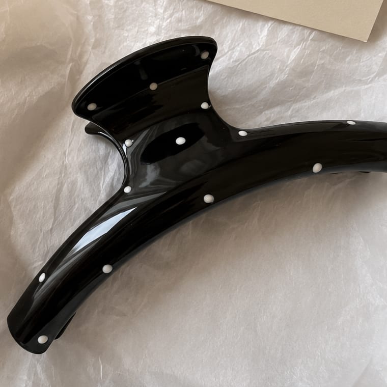 A glossy black hair claw clip decorated with small white pearl-like studs, resting on a textured white surface.