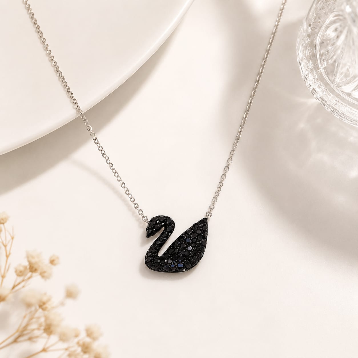 A silver chain necklace with a black swan pendant, styled on a white surface with a white plate, a glass, and dried flowers.