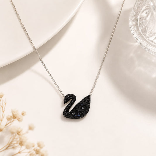 A silver chain necklace with a black swan pendant, styled on a white surface with a white plate, a glass, and dried flowers.