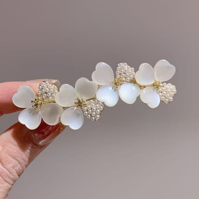 A close-up of a hand holding a decorative hair clip featuring translucent white flower petals, pearl-like bead clusters, and gold-tone centers against a soft gray background.