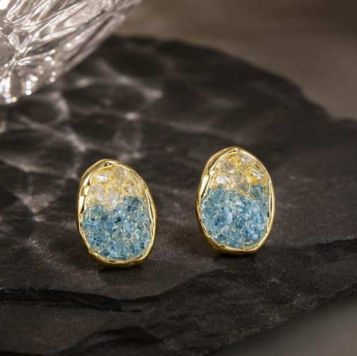 A pair of gold stud earrings with textured blue and yellow druzy stone inlays, displayed on a dark slate surface with a glass bowl in the background.