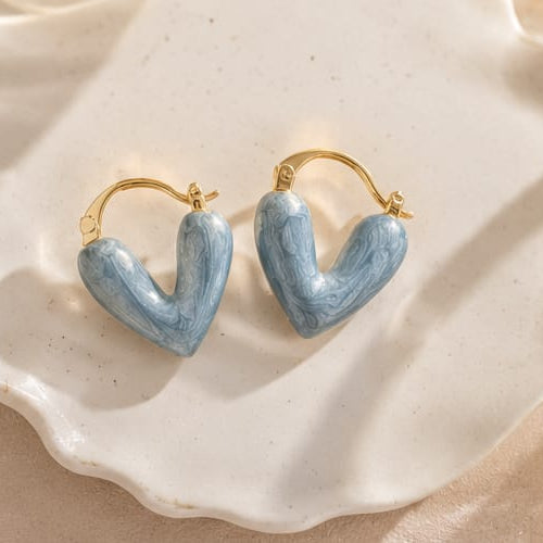 A pair of gold hoop earrings with light blue marbled heart-shaped drops, displayed on a scalloped white ceramic dish.