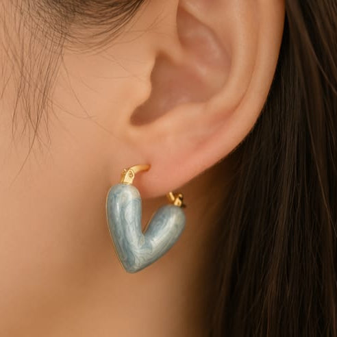 A close-up of a person's ear wearing a small heart-shaped hoop earring with a light blue marbled enamel face and gold-tone hoops.
