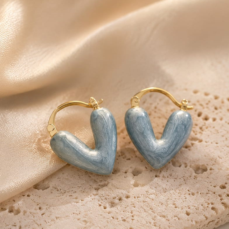 A pair of gold hoop earrings with light blue marbled heart-shaped charms, displayed on a textured beige stone slab with draped satin fabric in the background.