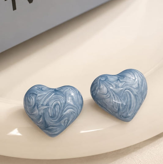 A pair of heart-shaped stud earrings with a light blue and white marbled swirl pattern, resting on a white ceramic dish. A gray box with the partial brand name 'TWAL' is visible in the top left corner.