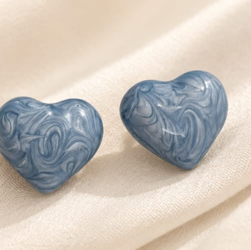 A pair of heart-shaped stud earrings with a blue and white marbled swirl pattern, displayed on a draped cream-colored fabric background.