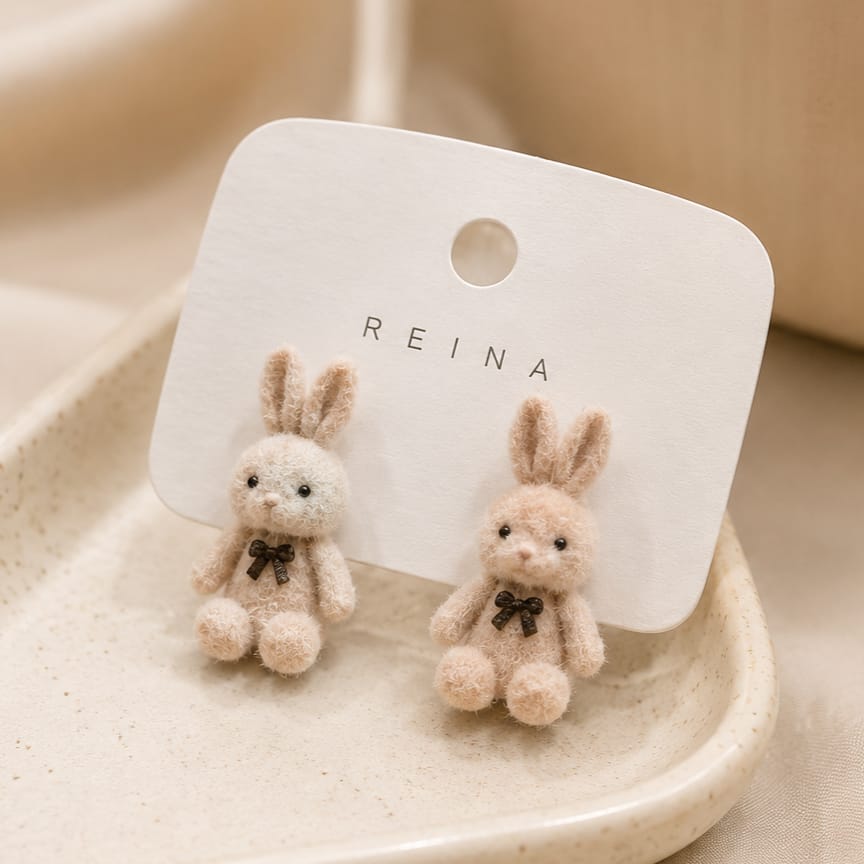 A pair of small bunny stud earrings with soft beige and white wool-like bodies and dark bow accents, displayed on a white REINA card holder in a warm neutral setting.