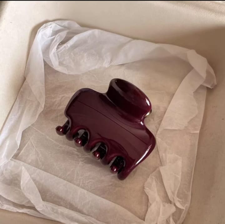 A glossy dark burgundy hair claw clip resting on white tissue paper inside a beige box.