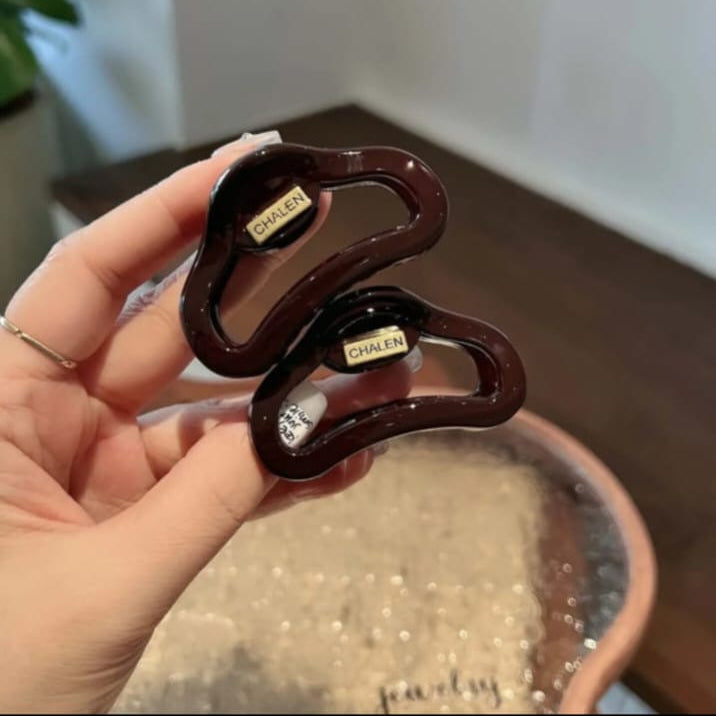 A hand holds two dark brown glossy hair claw clips with gold-tone metal accents labeled 'CHALEN' against a softly blurred indoor background.