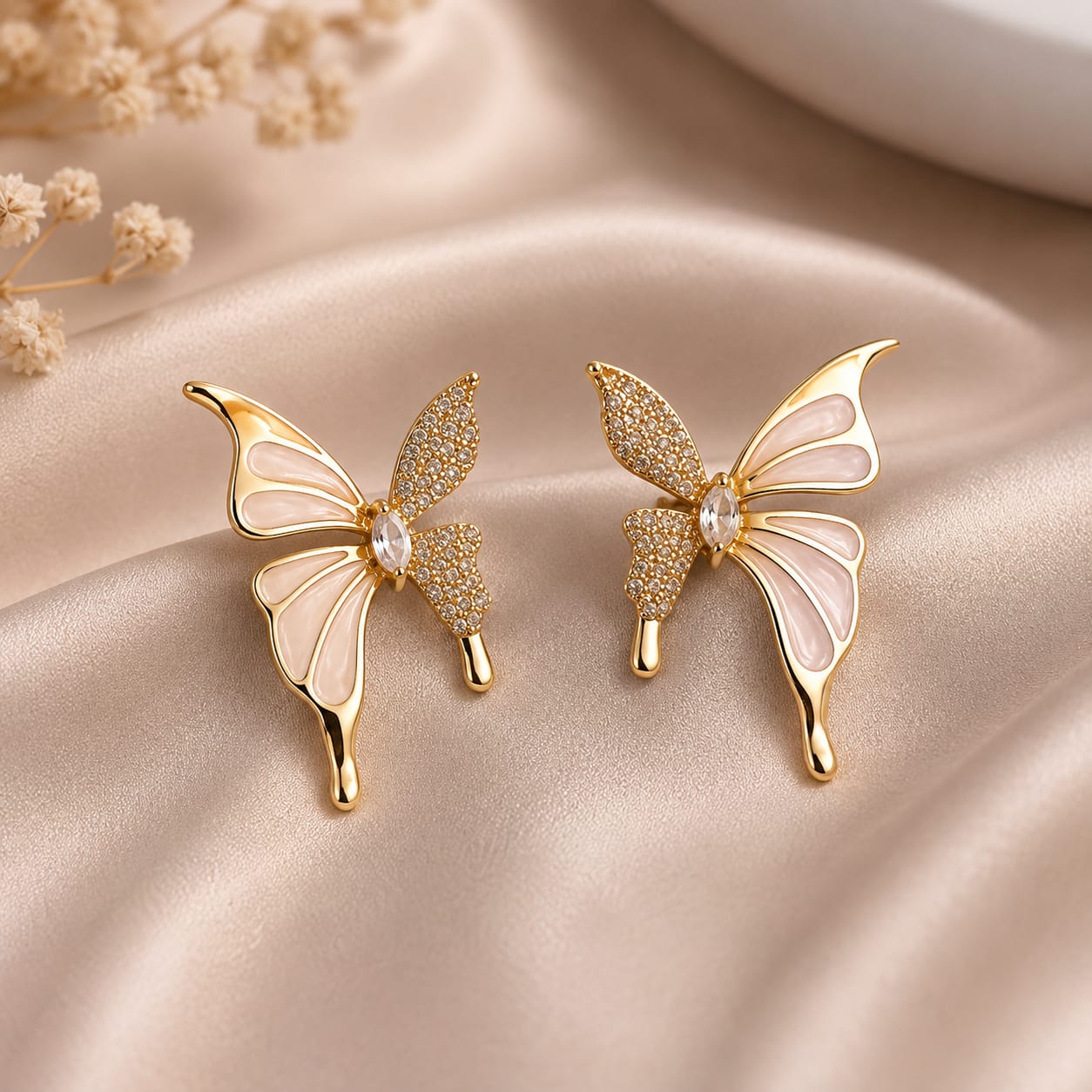 A pair of gold butterfly-shaped earrings with pale pink enamel wings and small clear stones, displayed on a beige satin fabric background with dried flowers.