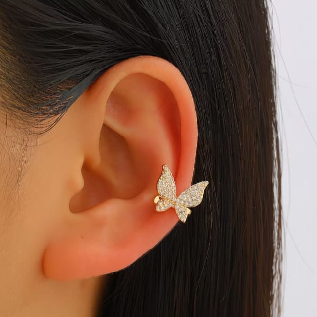 A close-up of a person's ear wearing a small gold butterfly stud earring adorned with clear pavé stones, set against dark hair and a light gray background.