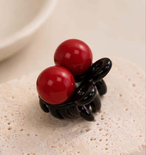 A glossy black claw-style hair clip with two large red spherical accents, photographed close up on a light textured surface.