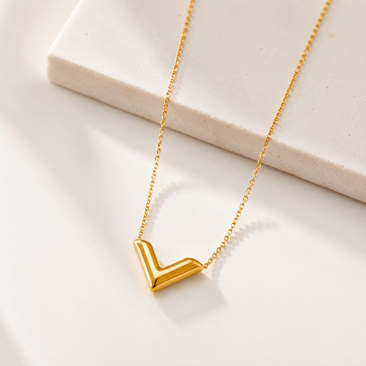 A gold necklace with a polished chevron V-shaped pendant, displayed on a clean white surface with a light beige stone block in the background.