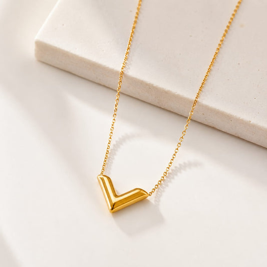 A gold necklace with a polished chevron V-shaped pendant, displayed on a clean white surface with a light beige stone block in the background.