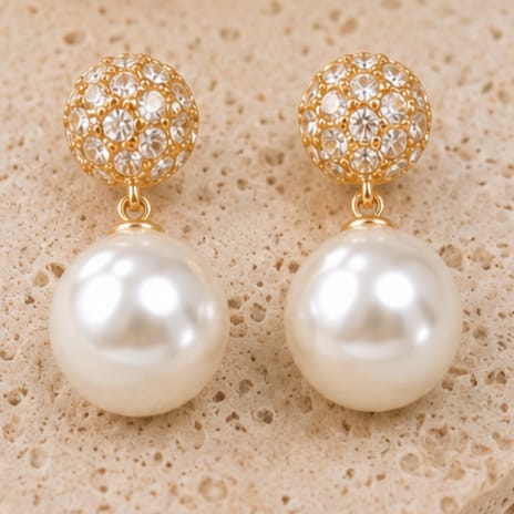 A pair of gold drop earrings with a crystal-encrusted spherical top and a large white pearl drop, displayed on a textured beige stone surface.
