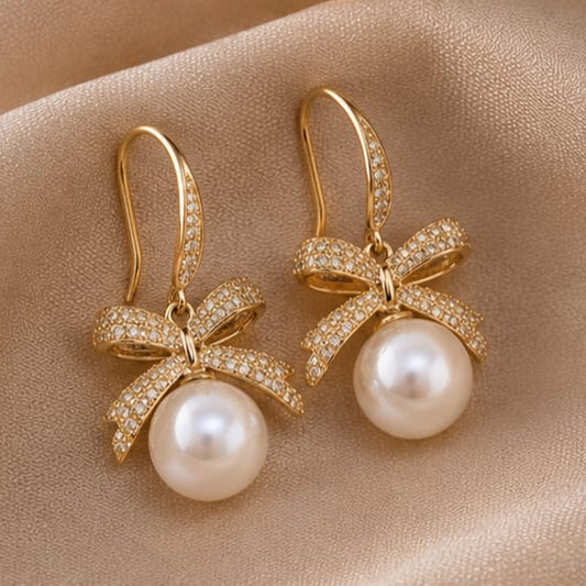 A pair of gold drop earrings with bow-shaped settings paved with small clear stones and a large white pearl hanging below, displayed on a beige satin fabric background.