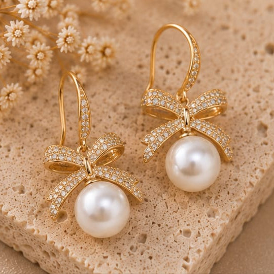 A pair of gold drop earrings with a bow-shaped setting paved with small clear stones and a large white pearl hanging below, displayed on a beige textured stone surface with dried flowers.