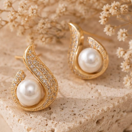 A pair of gold teardrop-shaped earrings with white freshwater pearls and pavé-set clear stones, displayed on a textured beige stone slab with dried baby's breath flowers.