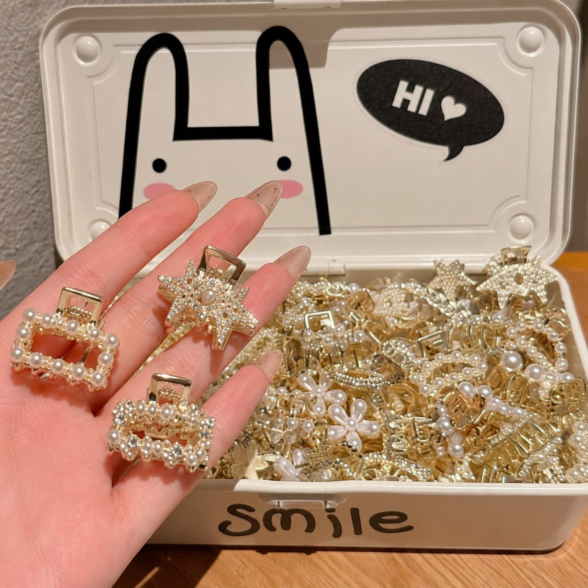 A hand holds three gold and pearl hair claw clips in front of an open white storage box filled with more clips. The box lid has a black bunny outline and a speech bubble that says 'HI', and the front says 'Smile'.