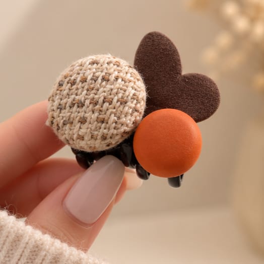 A close-up of a hand holding a decorative hair claw clip shaped like a ladybug. The clip features a beige and white houndstooth patterned body, a dark brown felt wing, a round orange body, and black spring claws.