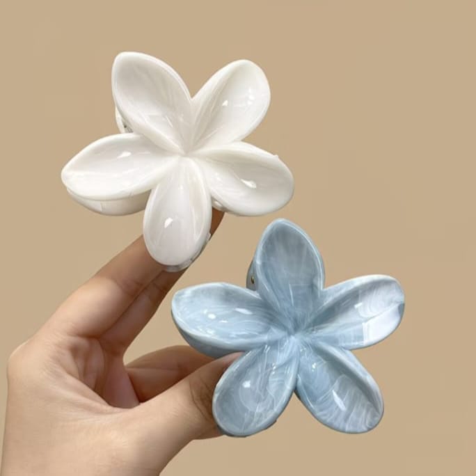A hand holds two glossy flower-shaped hair claw clips against a plain beige background. One clip is white and the other is light blue, both with a smooth, polished finish.