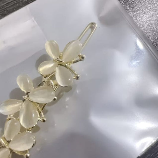A gold-tone hair claw clip decorated with a cluster of teardrop-shaped cream or off-white stone flowers, resting on a crinkled white plastic surface.