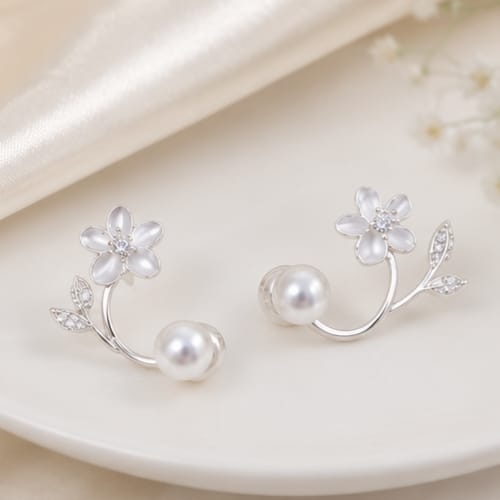 A pair of silver-toned ear cuffs styled as delicate flowers with white faux pearls and small clear rhinestones, resting on a warm cream-colored ceramic plate.