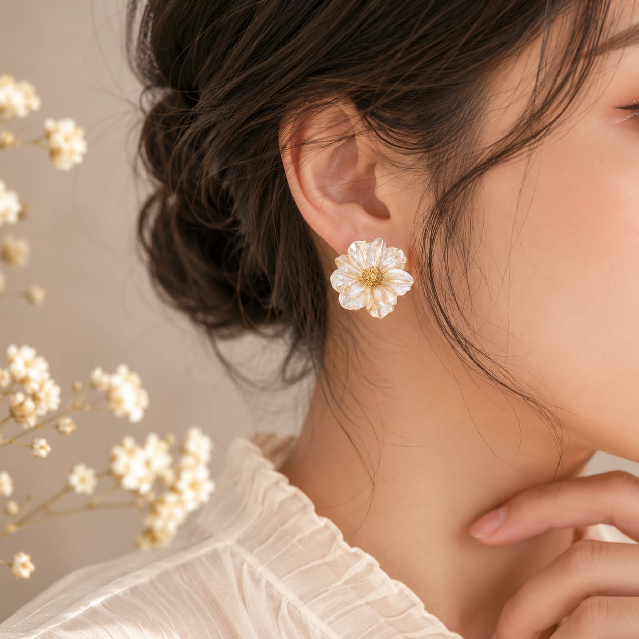 A close-up lifestyle shot of a woman wearing a delicate white flower stud earring with a gold center, styled with dried baby's breath flowers in the background.