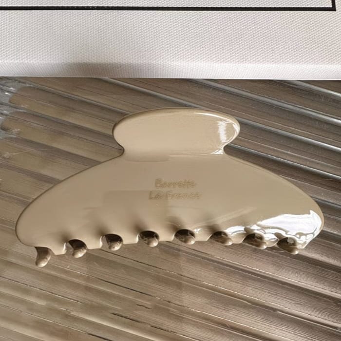 A glossy cream-colored hair claw clip with 'Berliet La France' printed on the front, resting on a metallic slatted surface.