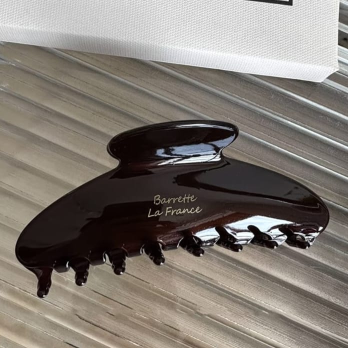 A glossy dark brown hair claw clip with the text 'Barrette La France' printed on it, resting on a metallic silver ribbed surface.