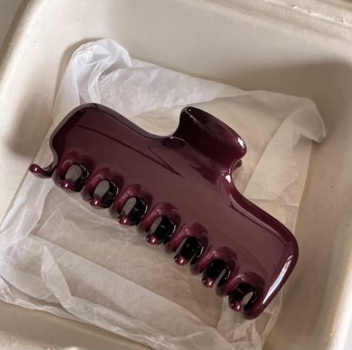 A glossy dark burgundy hair claw clip resting on white tissue paper inside a beige box.