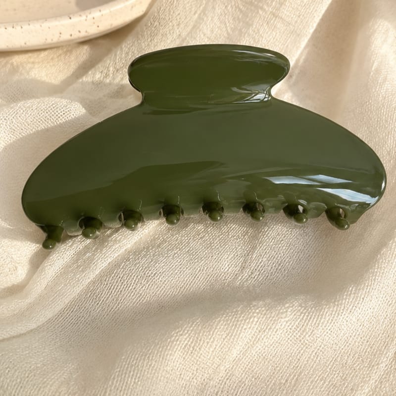 An olive green glossy hair claw clip resting on a textured cream fabric background.