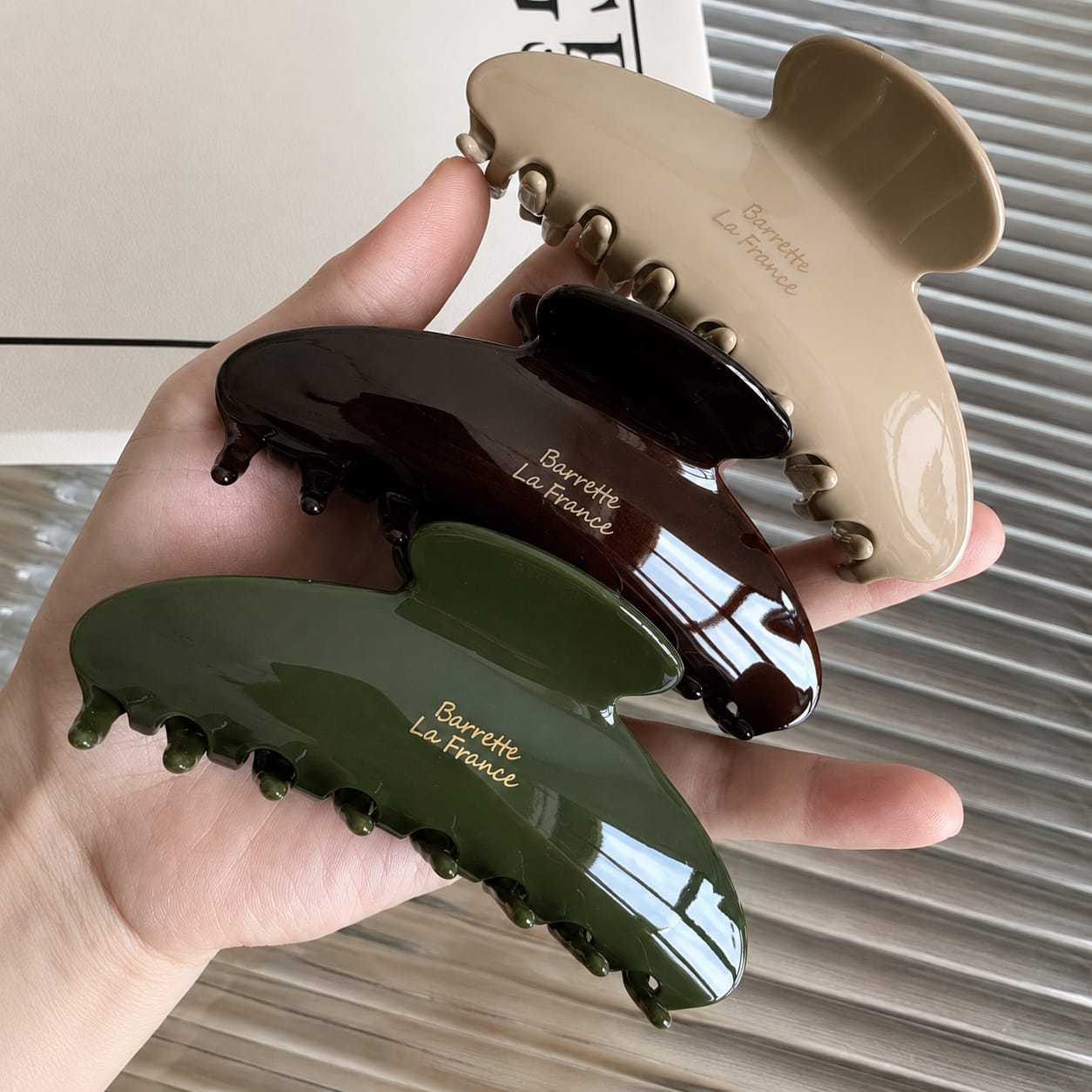 Three glossy hair claw clips in beige, dark brown, and olive green are held in a hand. Each clip has the text 'Barrette La France' printed on it.