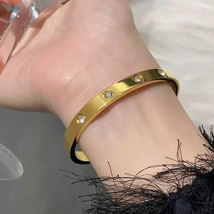 A gold bangle bracelet with small clear crystal accents, shown worn on a person's wrist against a soft light background.