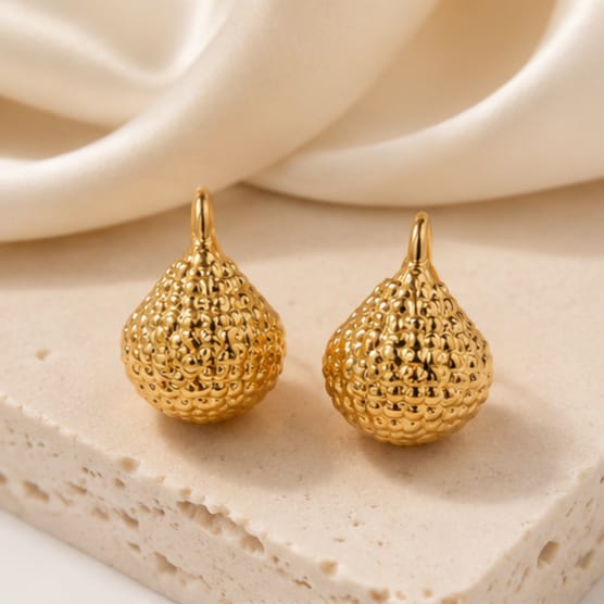 A pair of textured gold teardrop-shaped earrings with a small top loop, displayed on a light beige stone slab with a draped cream fabric background.