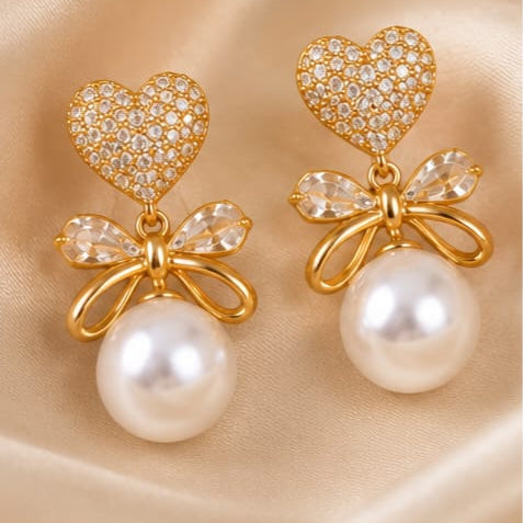 A pair of gold-tone drop earrings featuring a heart-shaped pavé crystal top, a clear crystal bow, and a large white pearl dangling below, displayed on a beige satin fabric background.