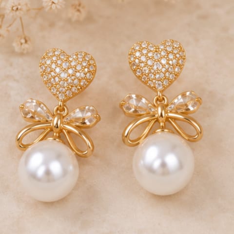 A pair of gold-toned drop earrings featuring a heart-shaped pavé top, a clear crystal bow motif, and a large white pearl dangling below, set against a soft beige background.